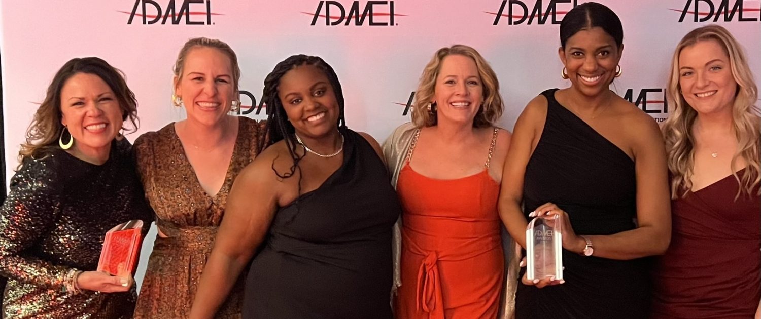 six women in evening gowns all in front of a white admei banner